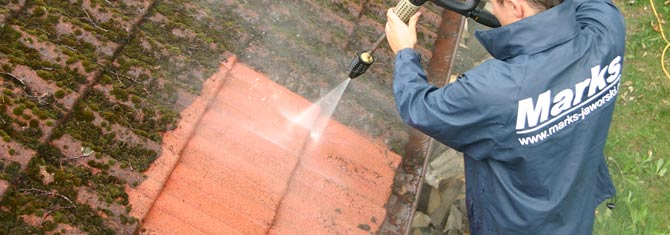 Специалист, при помощи аппарата высокого давления, смывает накопившийся мох на крыше дома